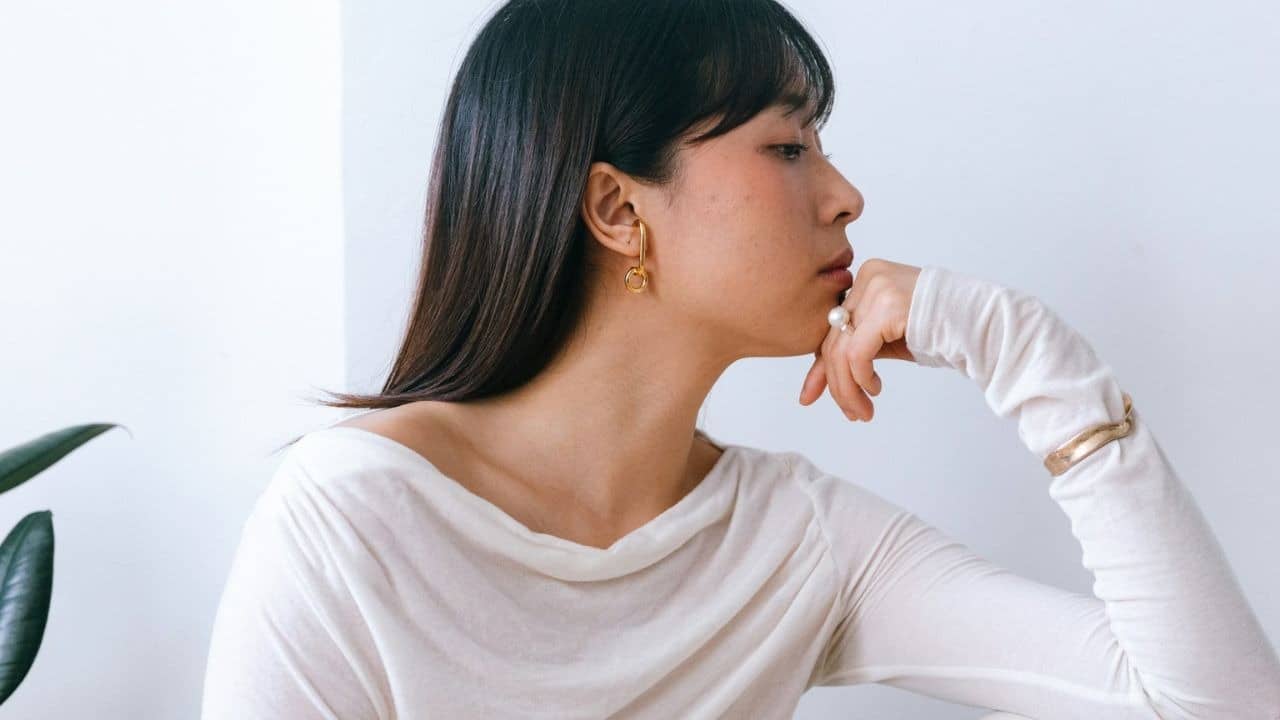 A dark haired lady sitting with her hand on her jaw, looking away from the camera.