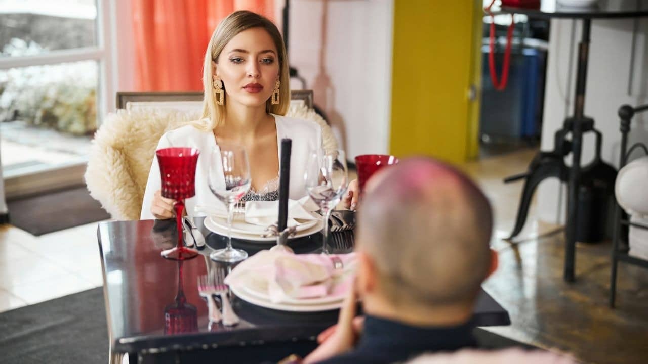 A woman with blonde hair sits at a restaurant table, holding a red glass.