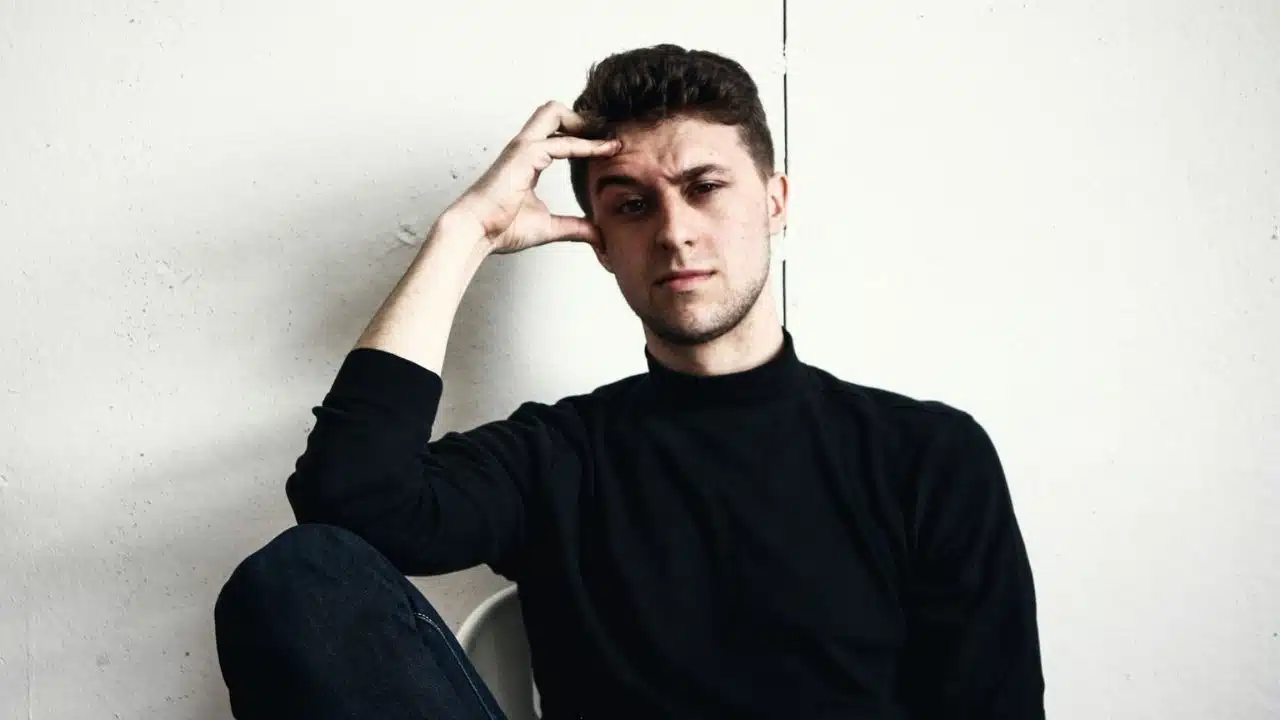 A man with black turtleneck is sitting with his hand on his head.