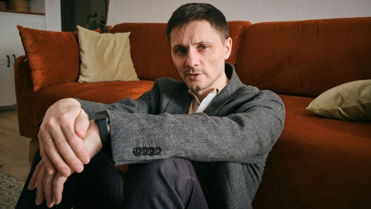 Man sitting on the floor, looking at the camera, in a tweed jacket.