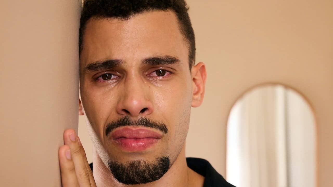 A man looking at the camera with tears in his eyes, standing by a wall.