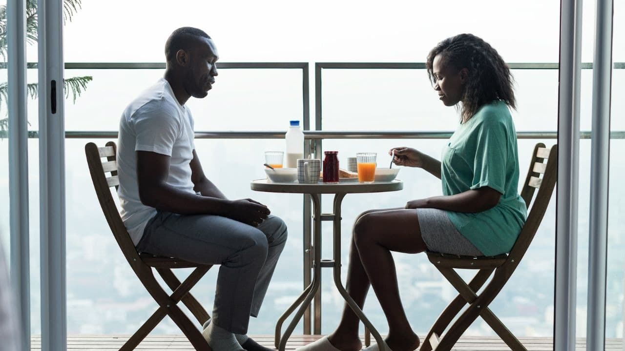 A couple sits at a small table on a balcony, looking at each other in silence.