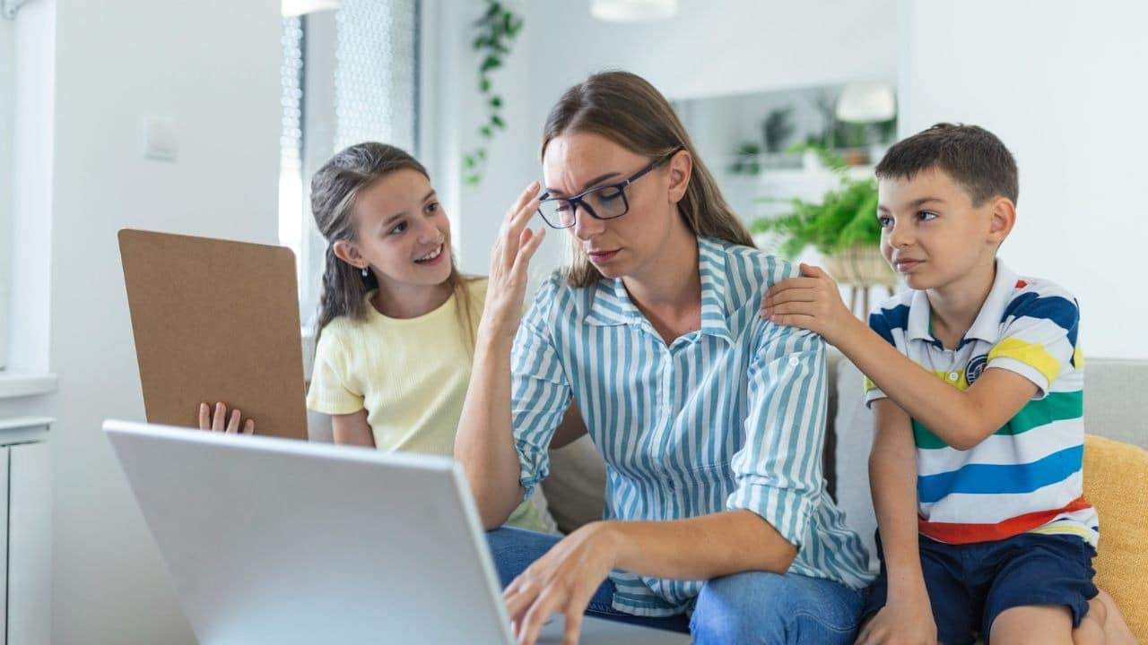 A stressed mother sits with her head in her hands while her two children look on.
