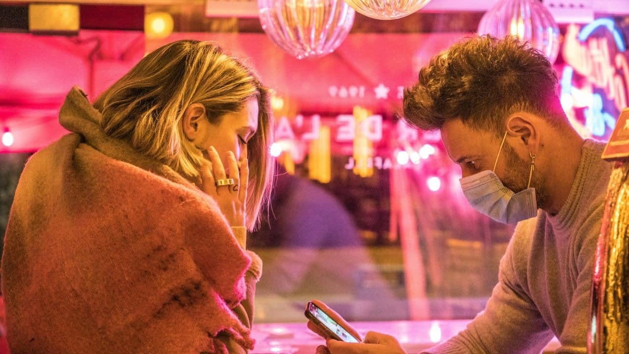 A man wearing a face mask looks at his phone while a woman looks at him.