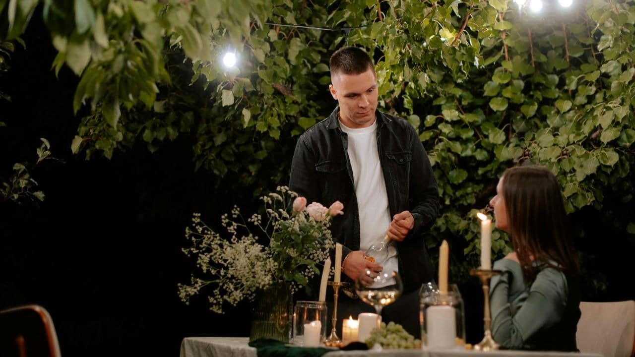 A man and woman sit at a table outdoors at night, with the man holding a wine bottle.