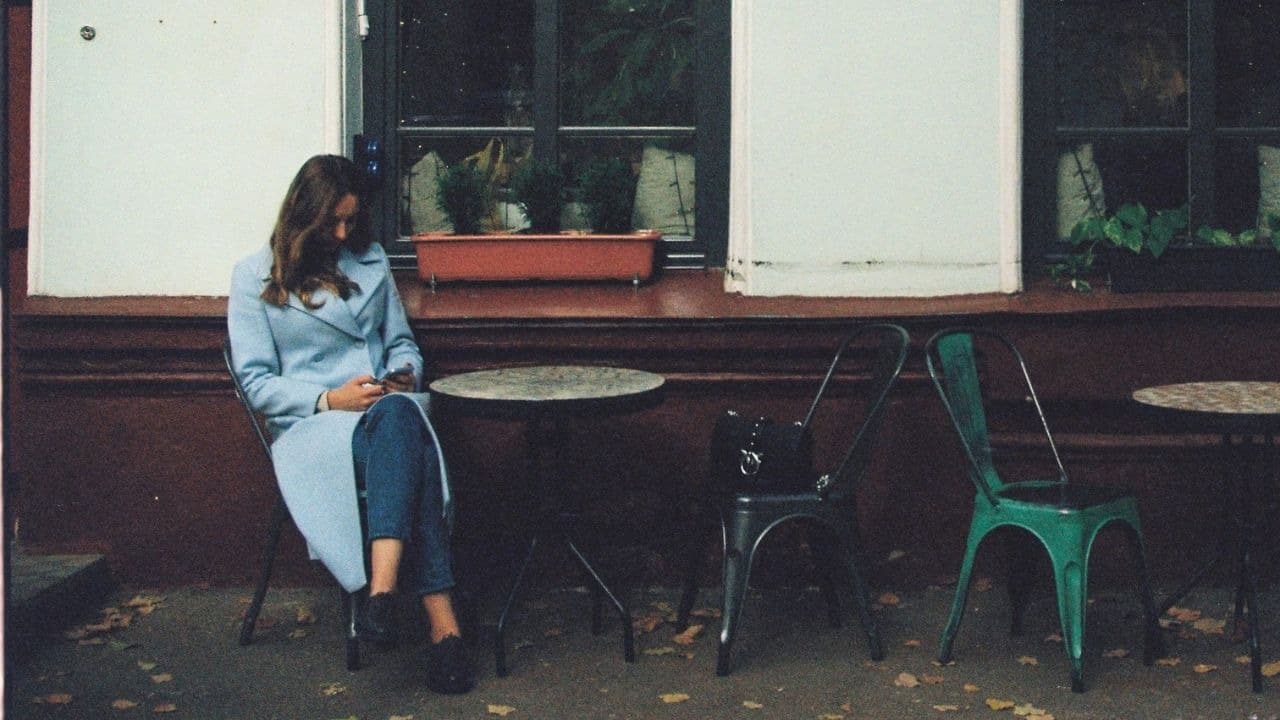 A woman in a light blue coat sits alone at an outdoor cafe table, looking at her phone.