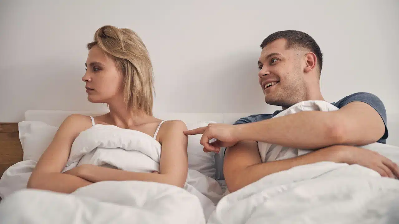 A smiling man poking a woman to get her attention as she looks away angrily while they lie together in a bed.