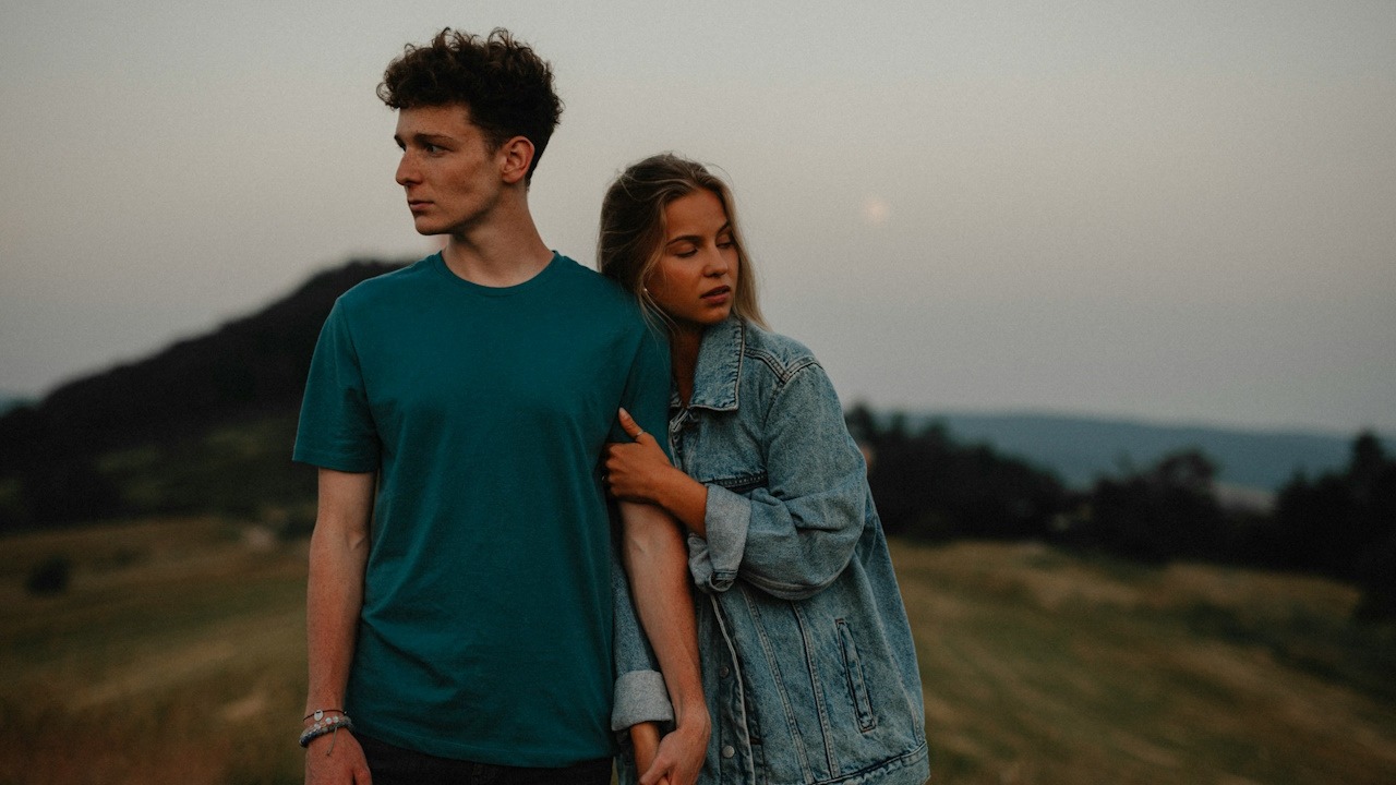 A young couple standing in nature in the countryside, holding hands but looking away from each other.
