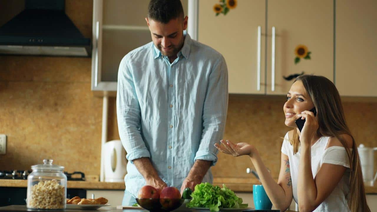 A woman is talking on the phone while her husband is preparing a meal.