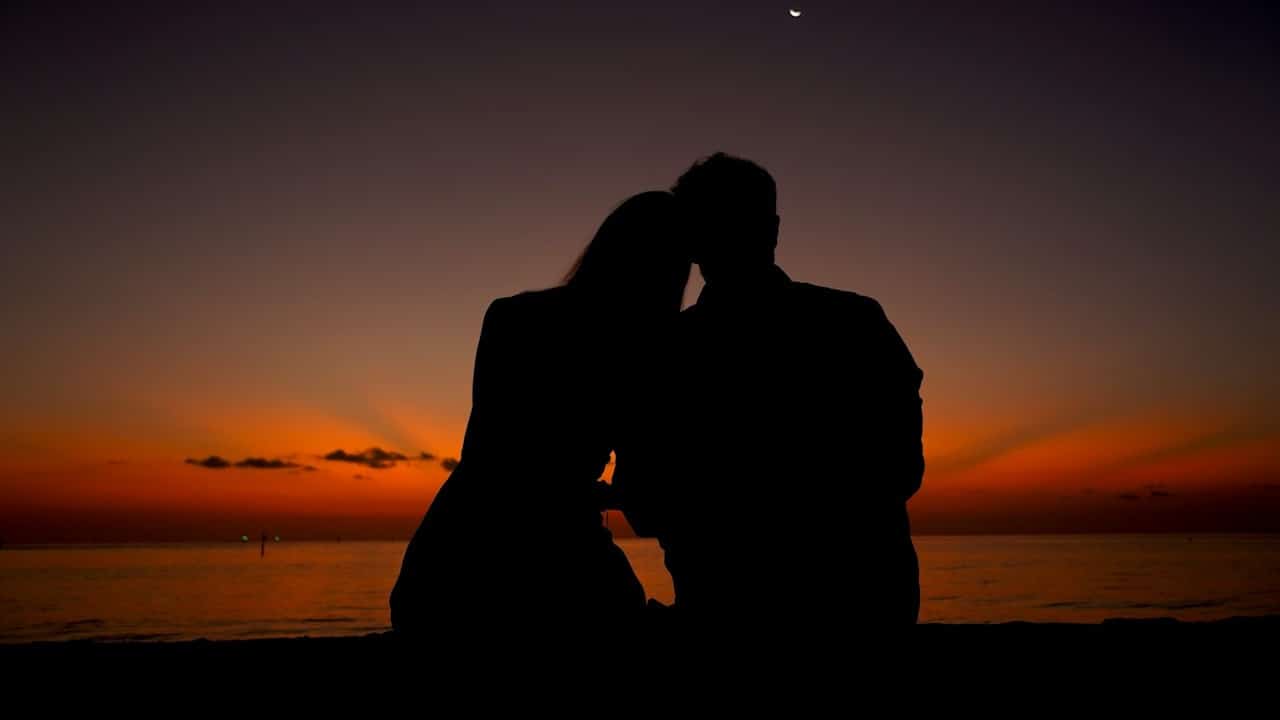 A silhouette of a couple sitting with their heads touching by the beach at sunset.