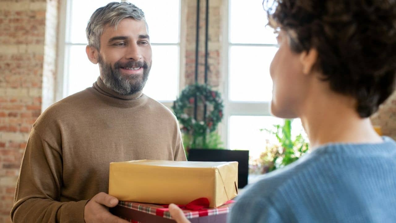 Smiling man with a gray beard and brown turtleneck holding wrapped gifts toward a woman with curly hair.