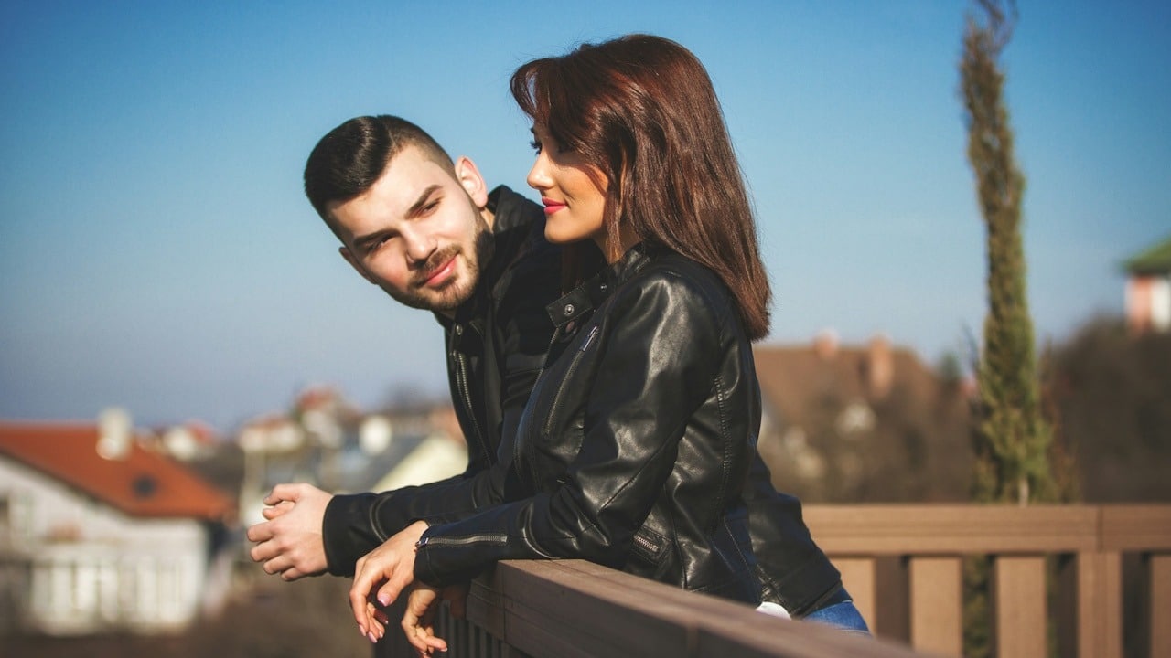 A man looking at a woman while they stand on a balcony overlooking an urban area.