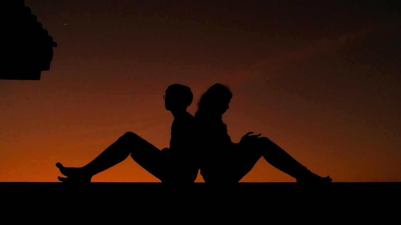 A couple standing together closely while standing on a bridge with the sun in the back.