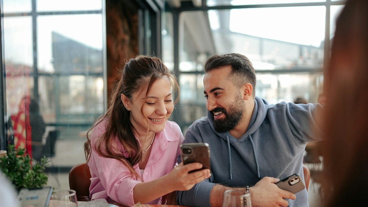 A woman smiling and showing something on her phone to a bearded man who’s also smiling.
