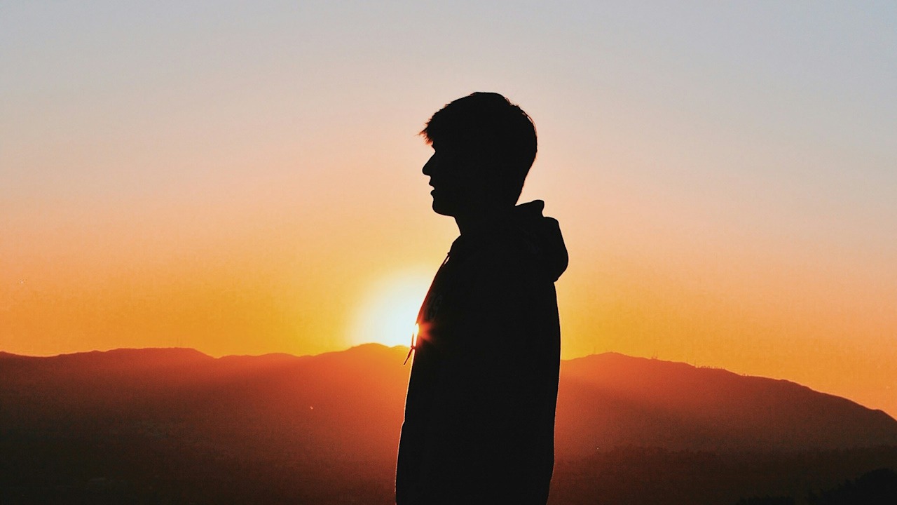 A man standing outdoors at sunset time with mountains in the background.