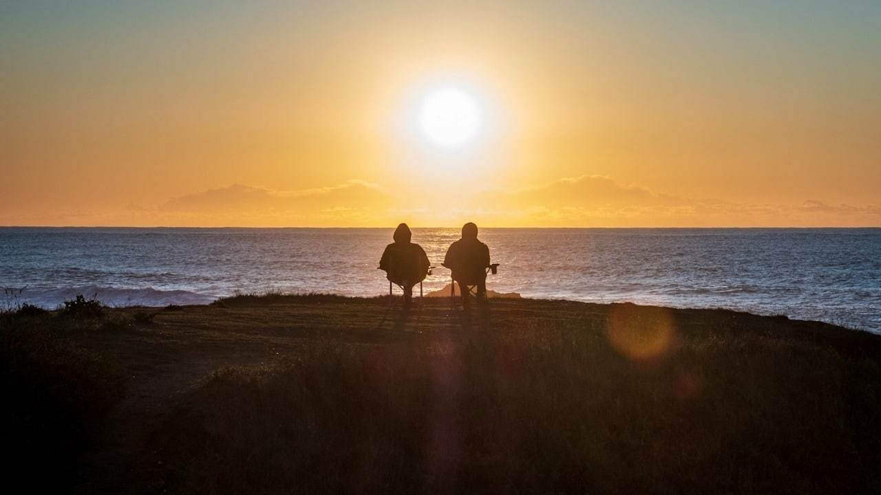A couple sitting on a cliff overlooking the ocean with the sun in the sky.