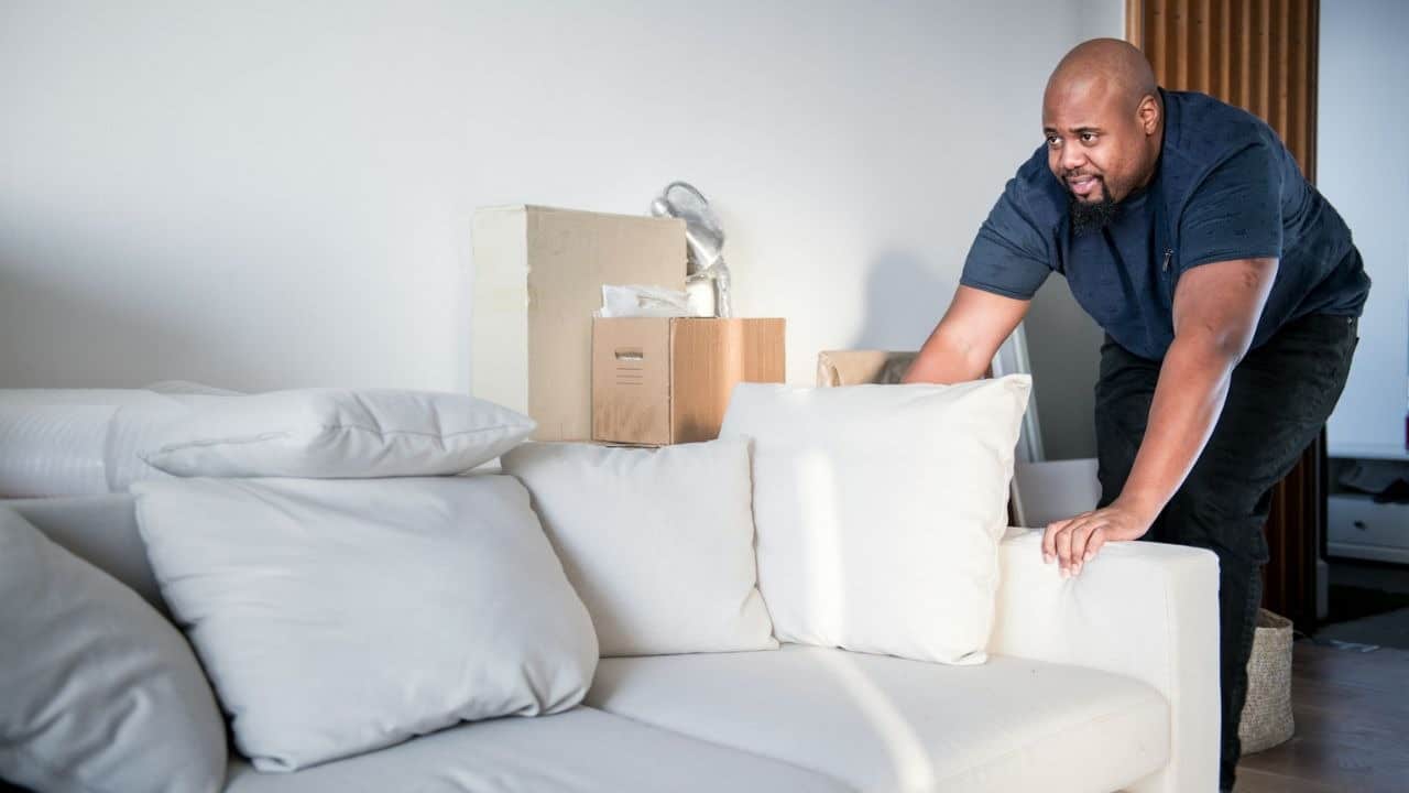 A black man is pushing a white sofa from behind with his hand on the armrest. The sofa's back has cardboard boxes.