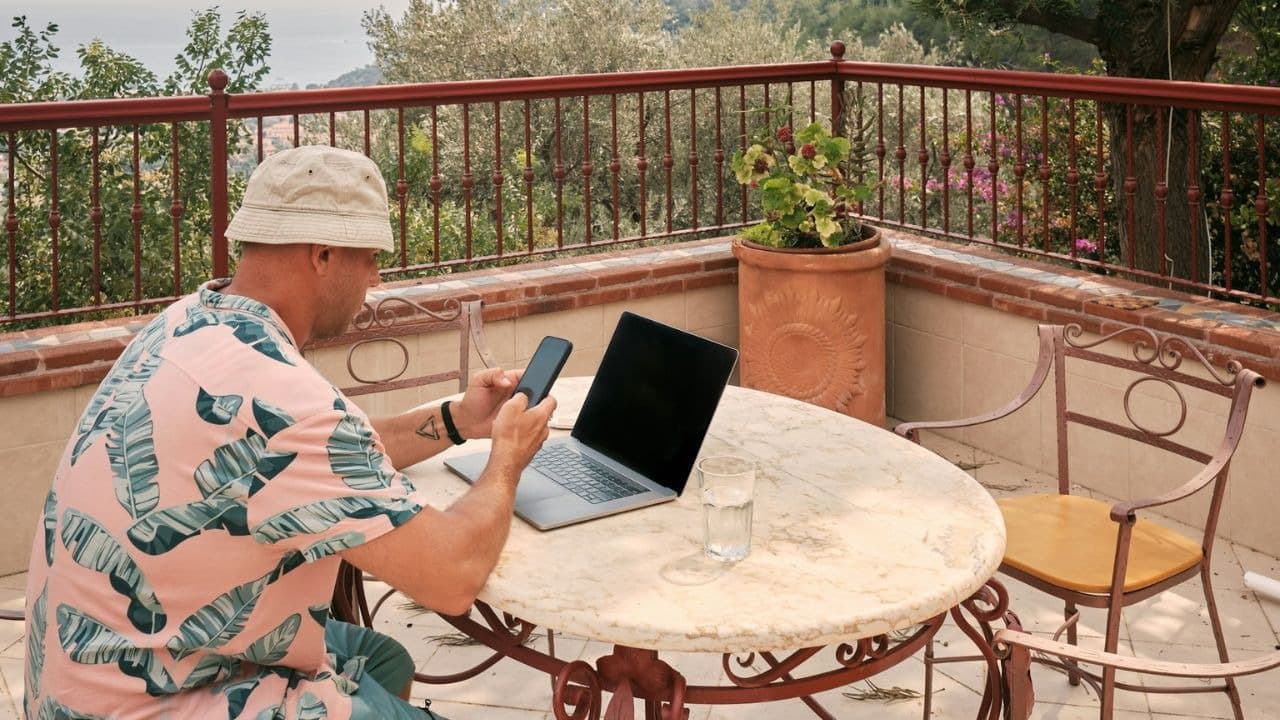 A man is reading a news article on his phone and has his laptop while standing on a balcony.