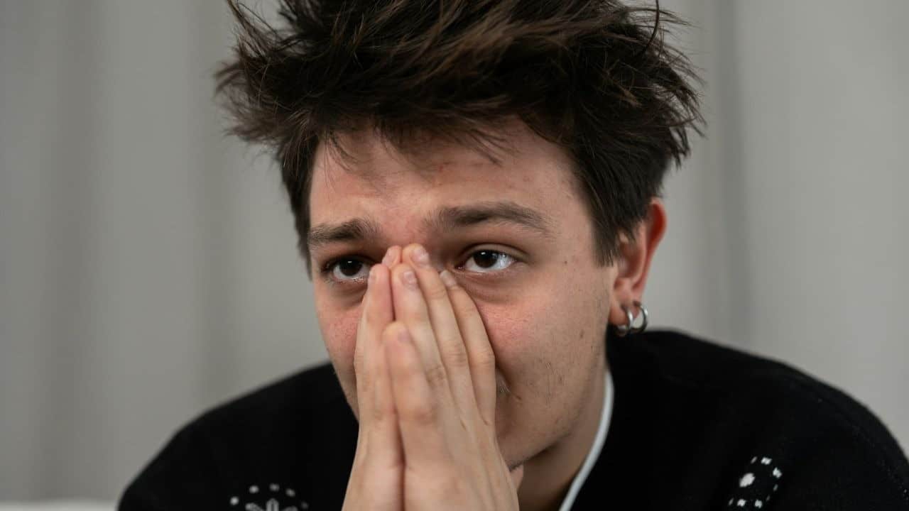 A man with brown hair and an earring on his left ear is sitting with his hands over his face.