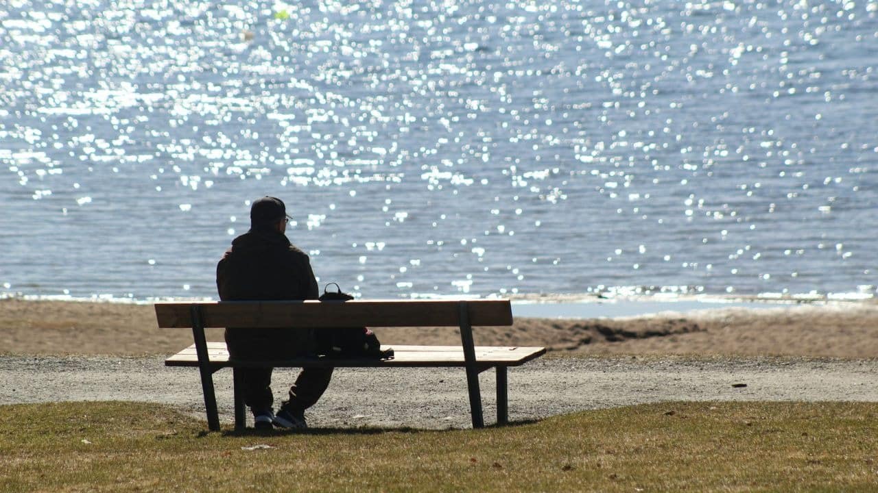 A man is sitting on a bench, wearing a hat and jacket, with the sun shining on the water.