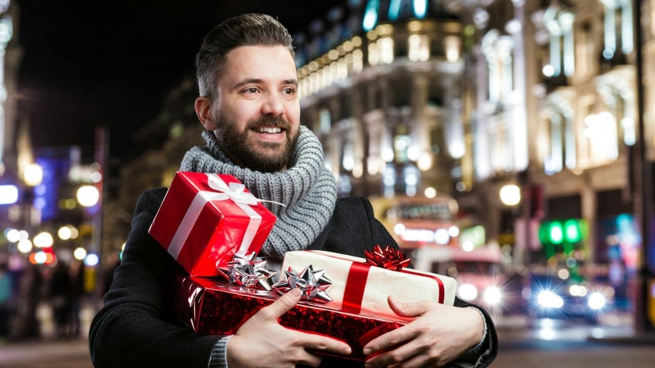 A man with a beard and a gray scarf is standing on a city street at night, holding several wrapped gifts.