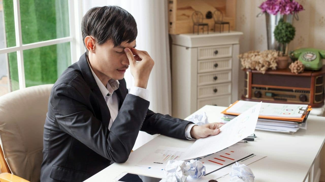 A man in a suit jacket is sitting at a desk with his hand on his eyes, looking at papers.