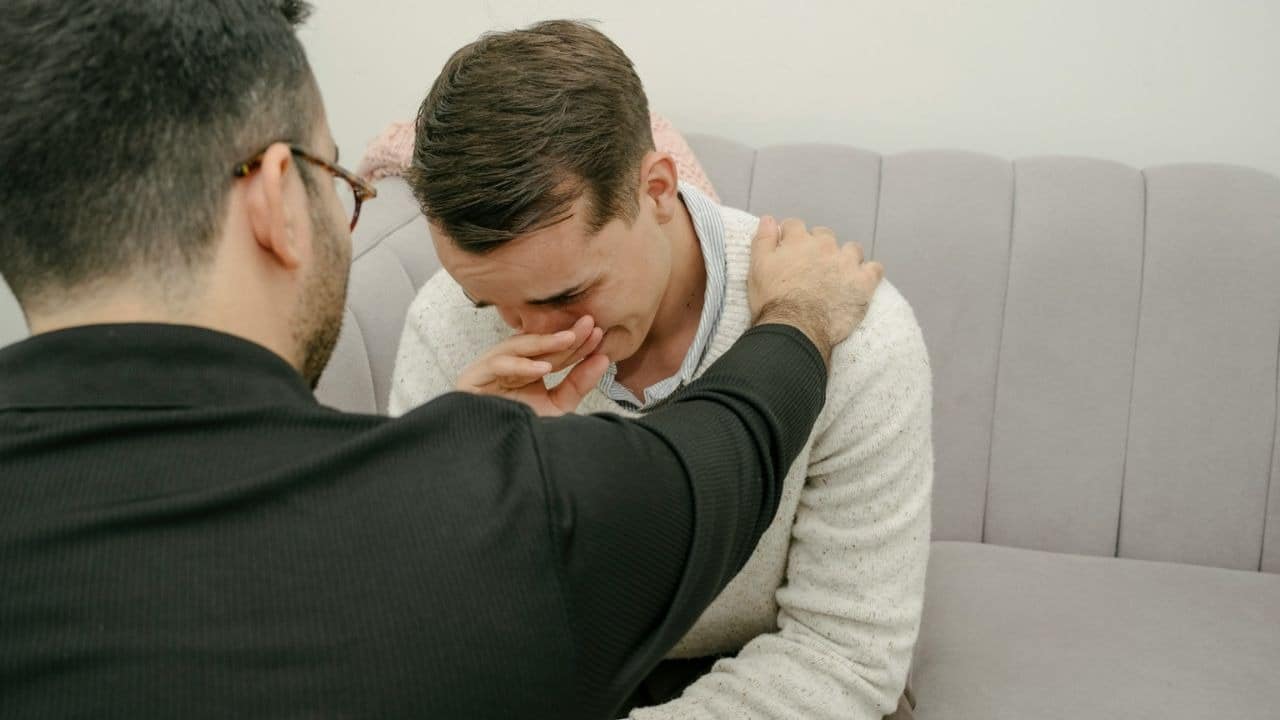 A man wearing a white collared shirt with his hand on his forehead is looking at another man.