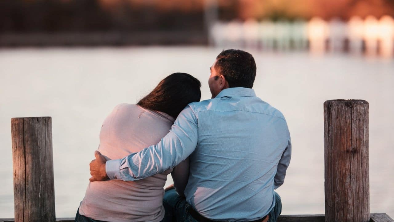 A pair of people is sitting on a wooden bridge overlooking a body of water, holding hands and gazing at the water.