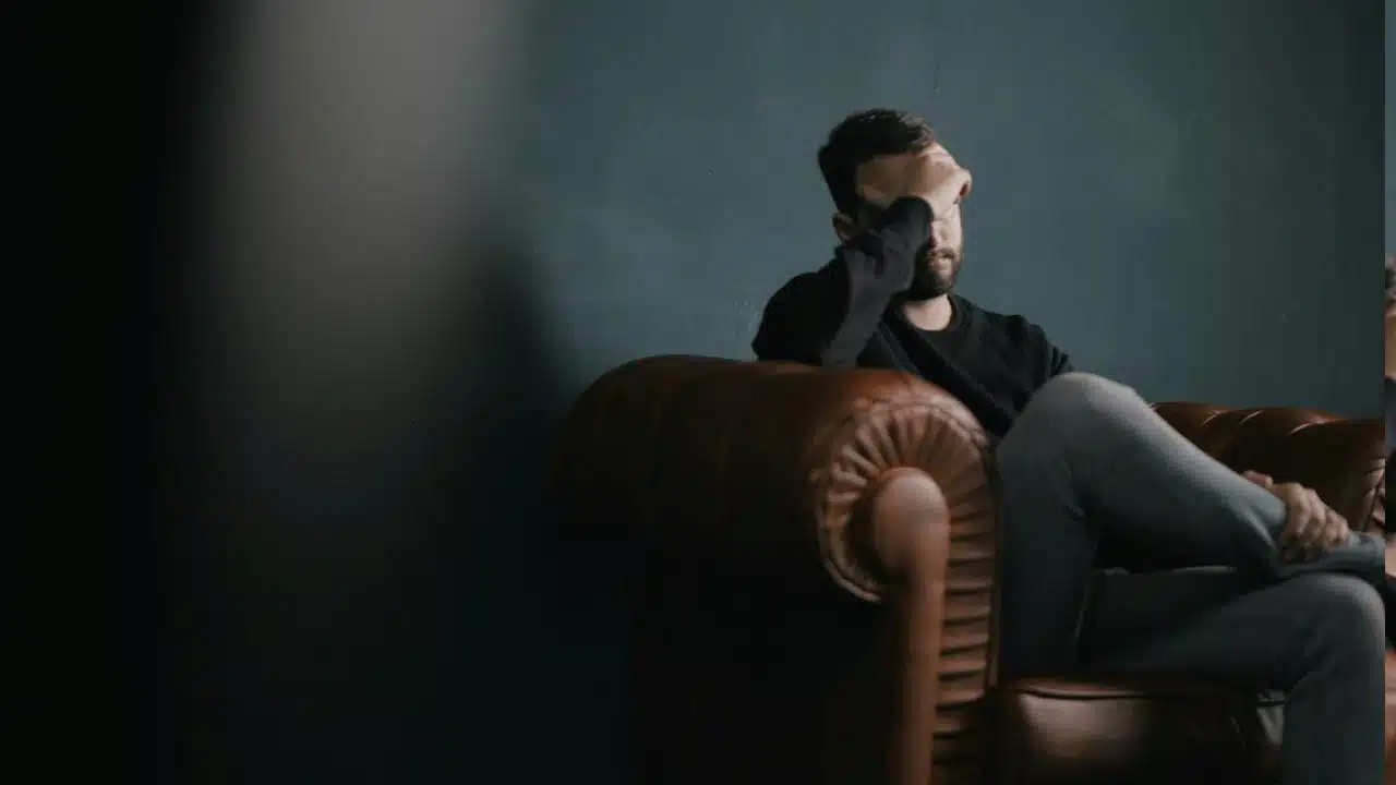 A depressed man holding his head with his hand while sitting on a sofa.