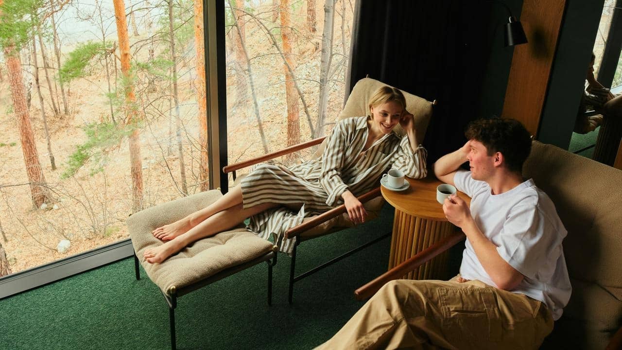 A couple relaxing with coffee by a large window overlooking trees.