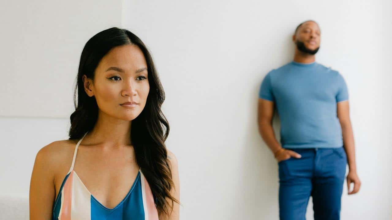 A woman standing in the forefront while a bearded man leans against a wall in the background.