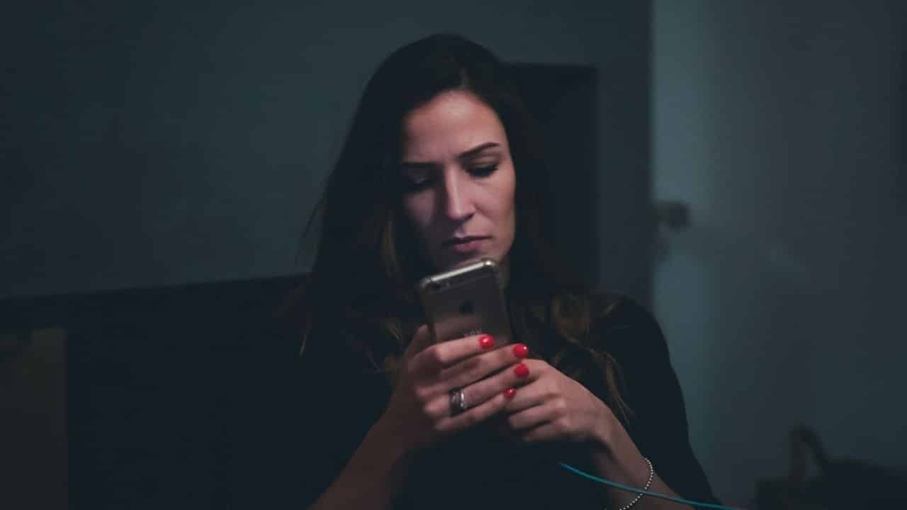 A woman with red nail polish looks intently at her phone in a dimly lit room.