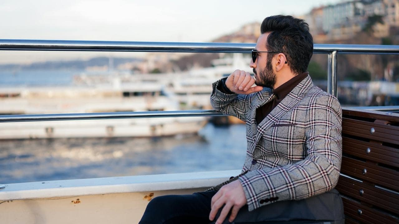 A man in a plaid blazer and sunglasses sitting by the water.