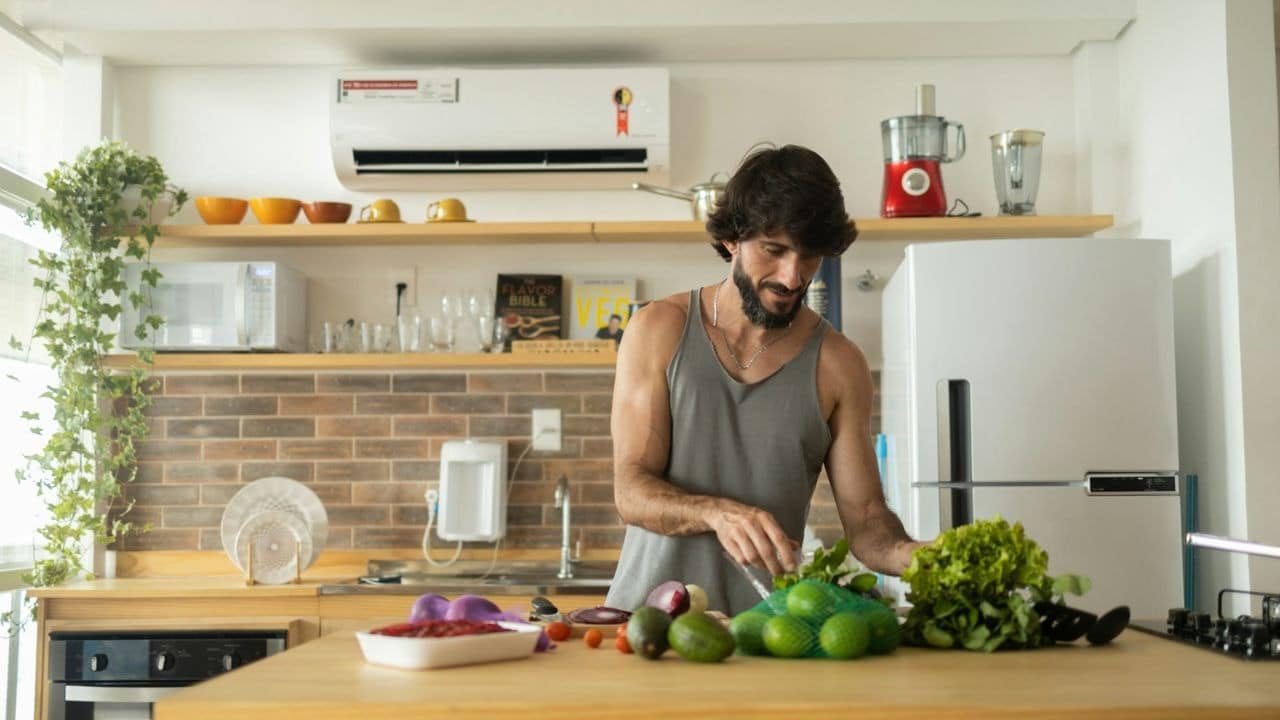 Bearded man in a tank top preparing fresh vegetables in a brightly lit kitchen.