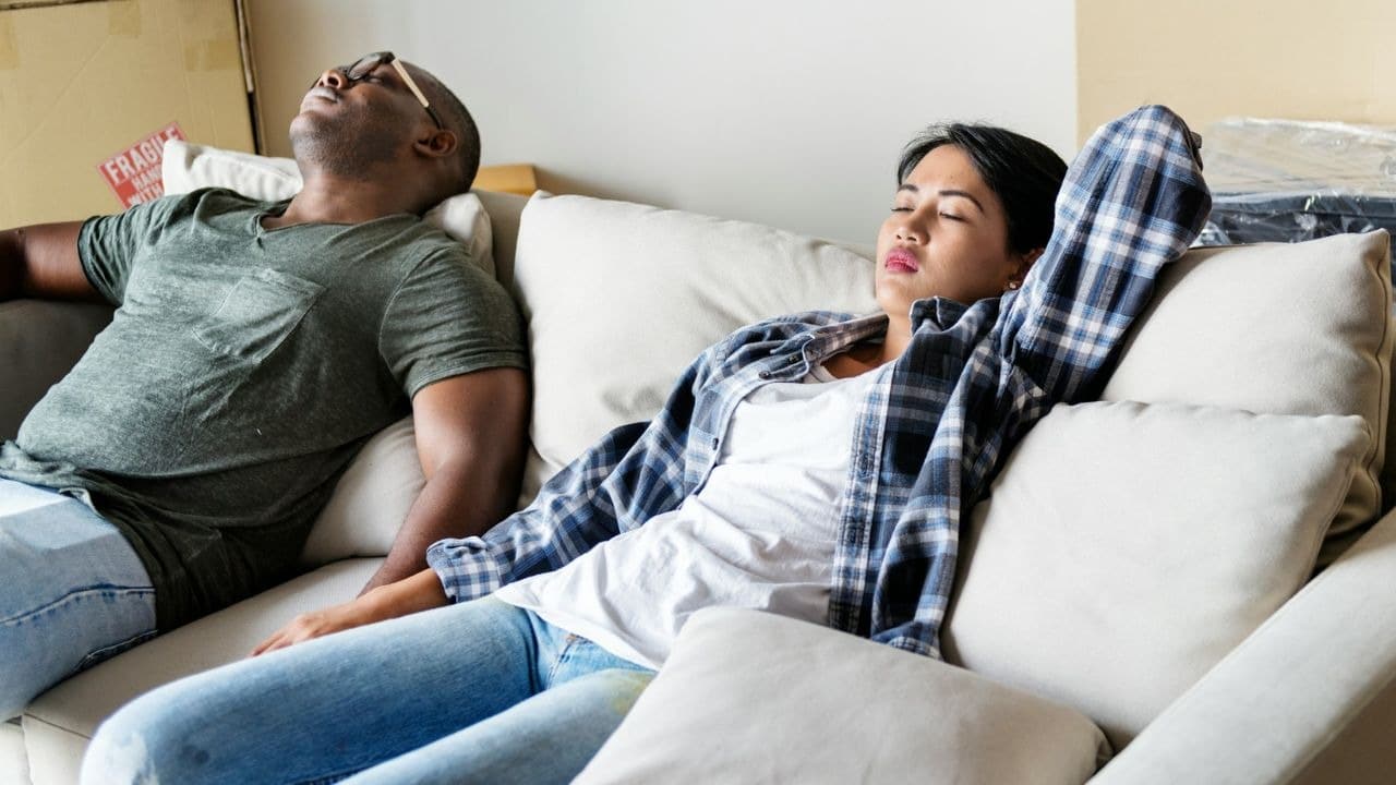 A tired man and woman sleeping on a couch in a messy living room.