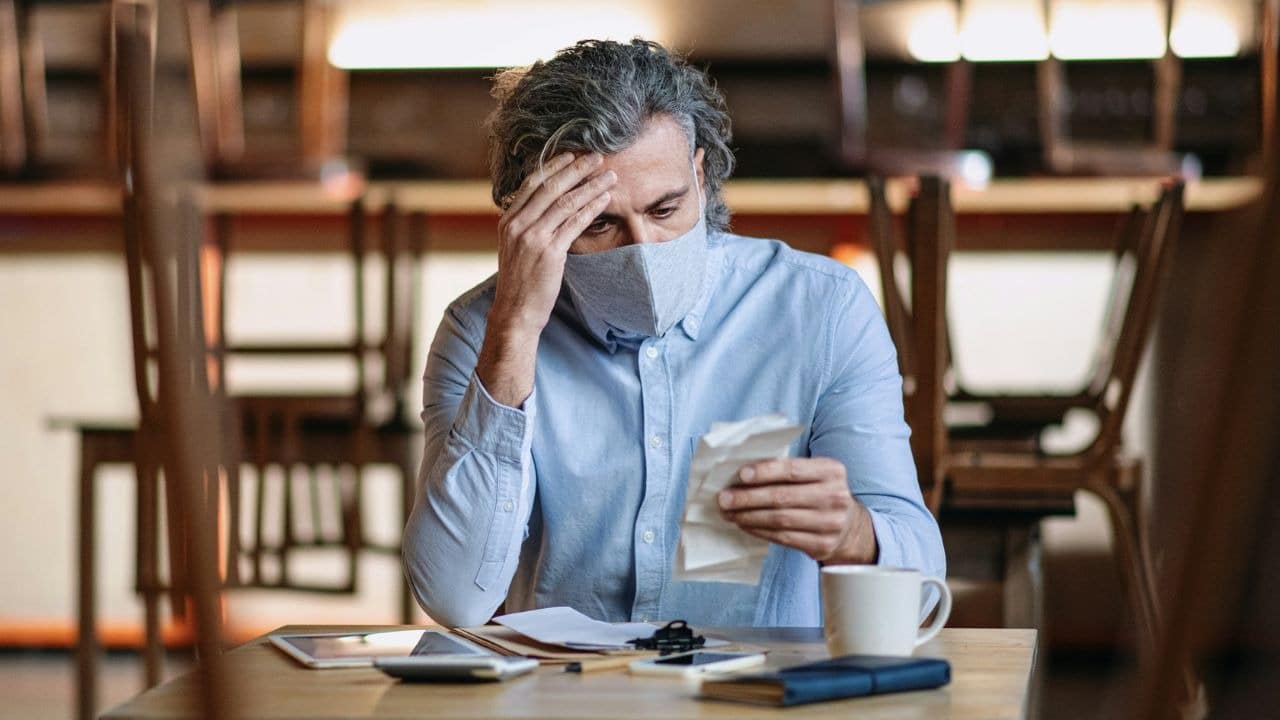 A frustrated man wearing a face mask looks at a long receipt with his head in his hand.