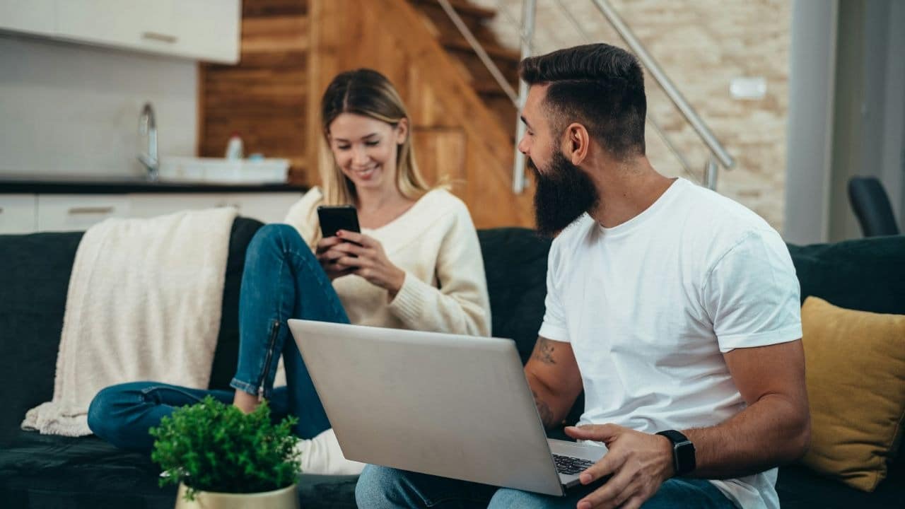 A smiling couple sits on a couch, each looking at their own device, a phone and a laptop.