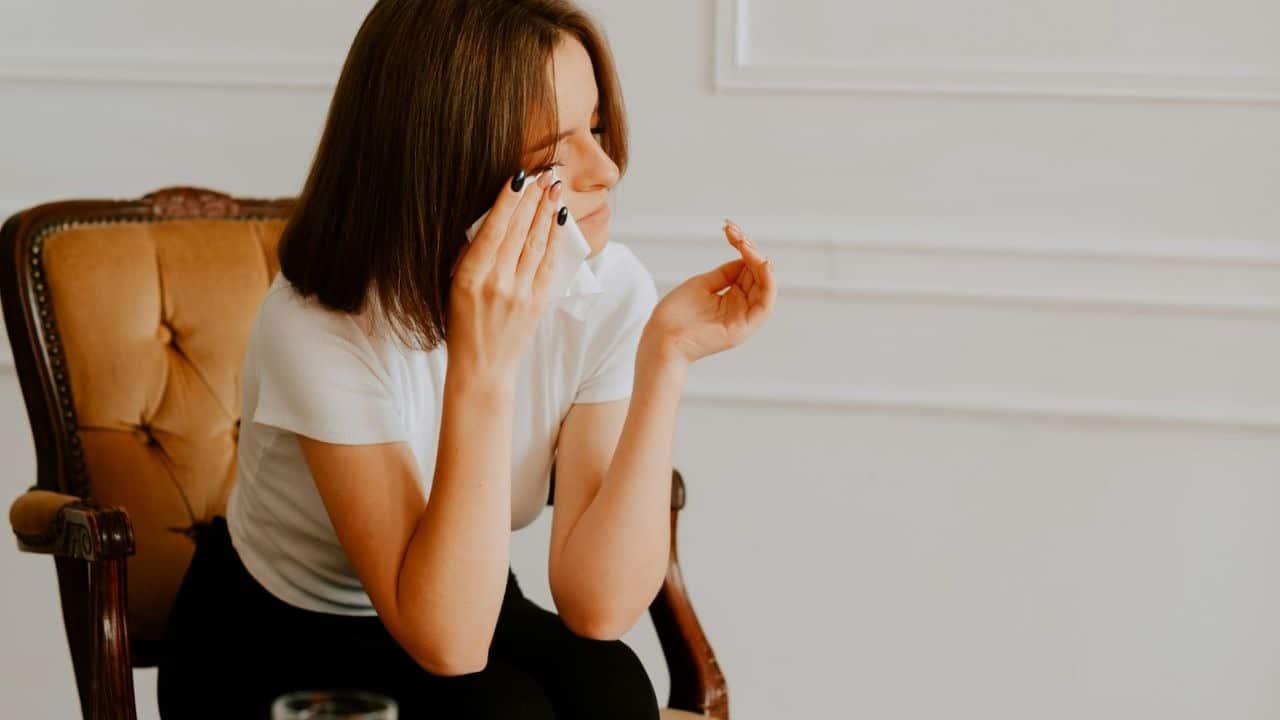 A woman in a white shirt sits in an armchair, wiping her eyes with a tissue.