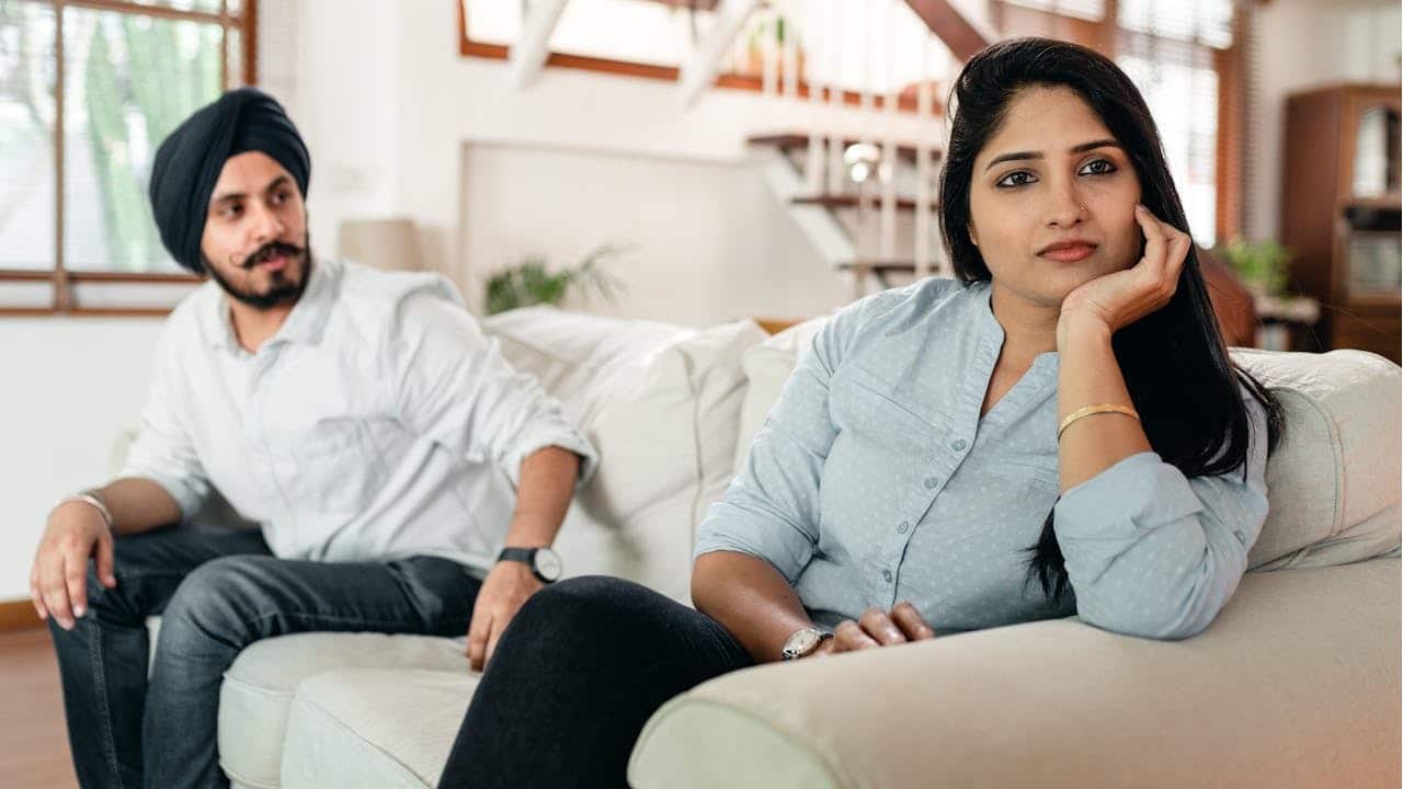 A woman sits on a couch while a man in a turban sits nearby looking at her with a concerned expression.