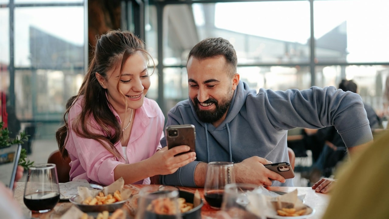 A woman showing something to his husband on her phone while they are having a meal together in a restaurant.