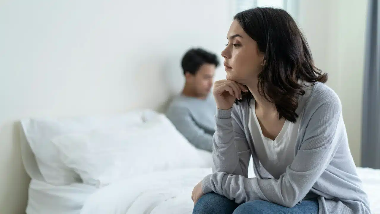 A woman sitting on a bed and thinking while a man sits in the background.