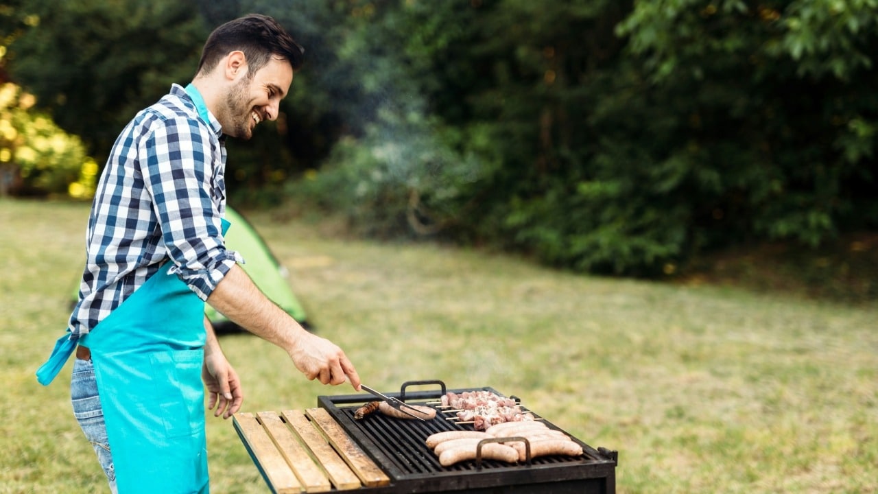 A man wearing a blue apron smiles while cooking sausages and meat on an outdoor grill.
