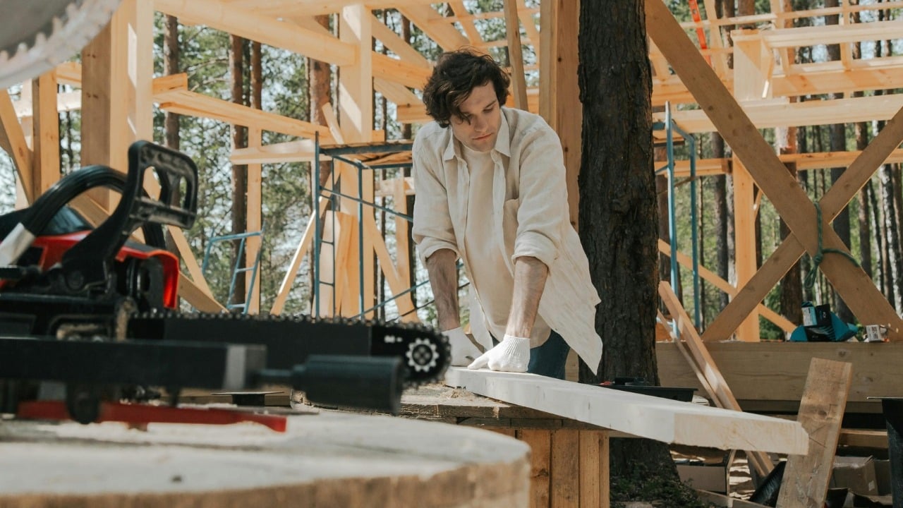 A man in a white shirt and gloves works on a wooden beam at a construction site.