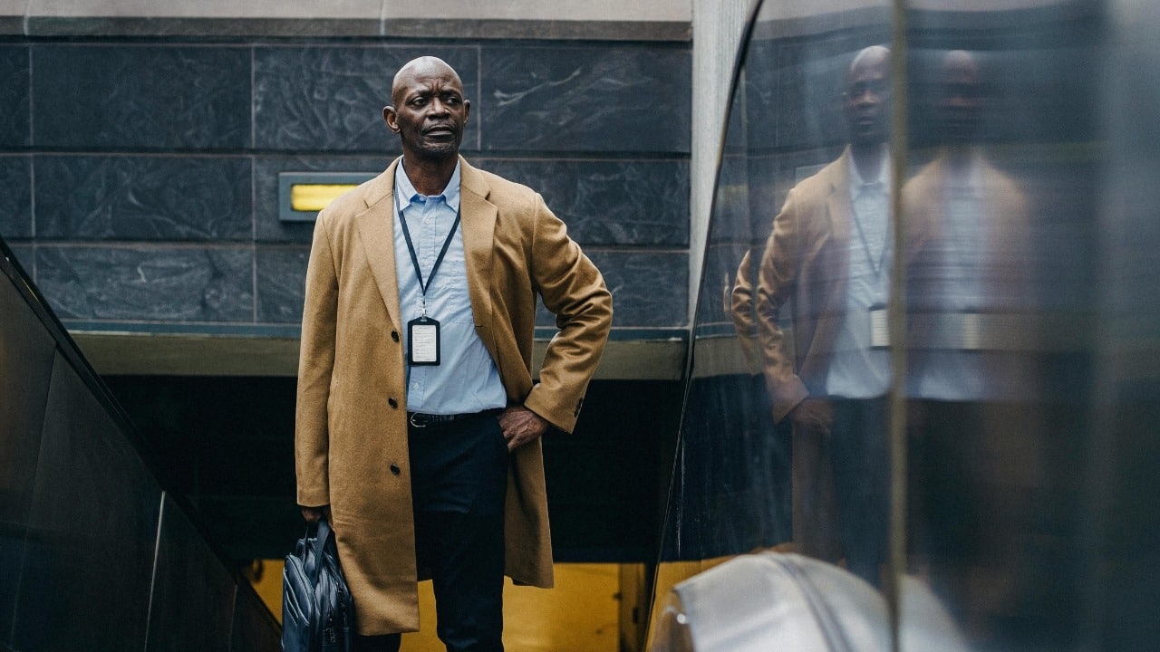 A man in a tan coat and blue shirt stands near an escalator in a building.