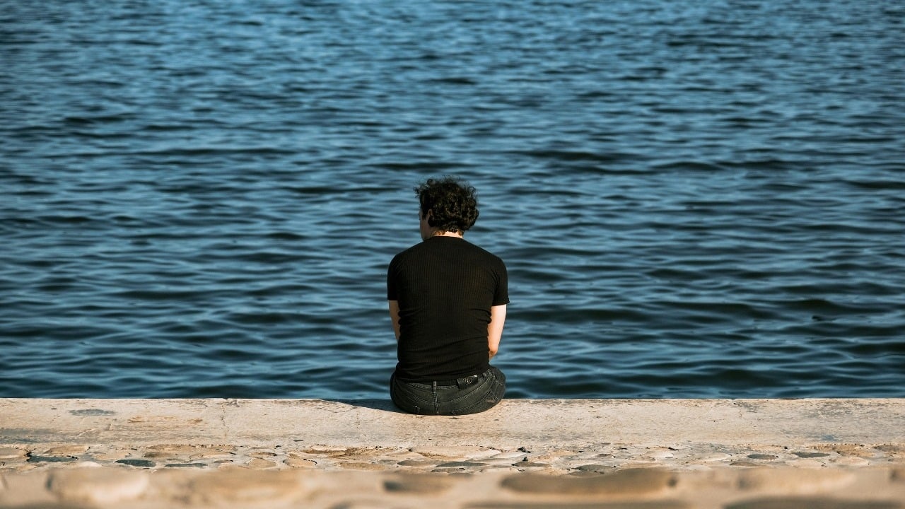 A person with dark curly hair sits on a stone ledge facing a large body of water.
