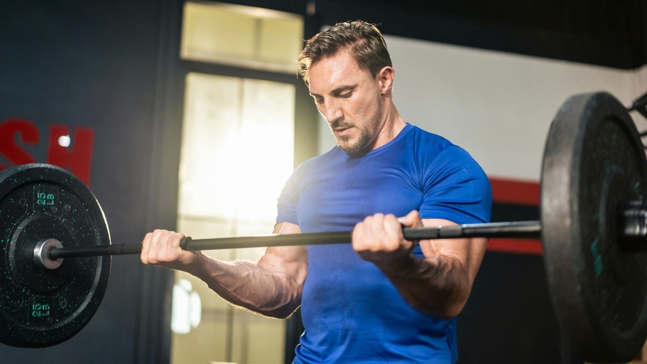 A muscular man in a blue shirt lifts a weighted barbell during a workout indoors.
