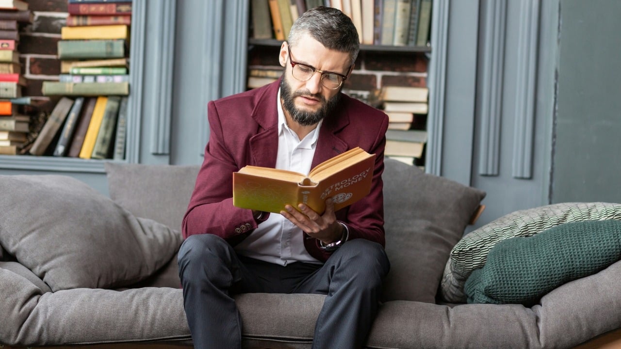 A man in a maroon blazer sits on a sofa while reading an orange book.