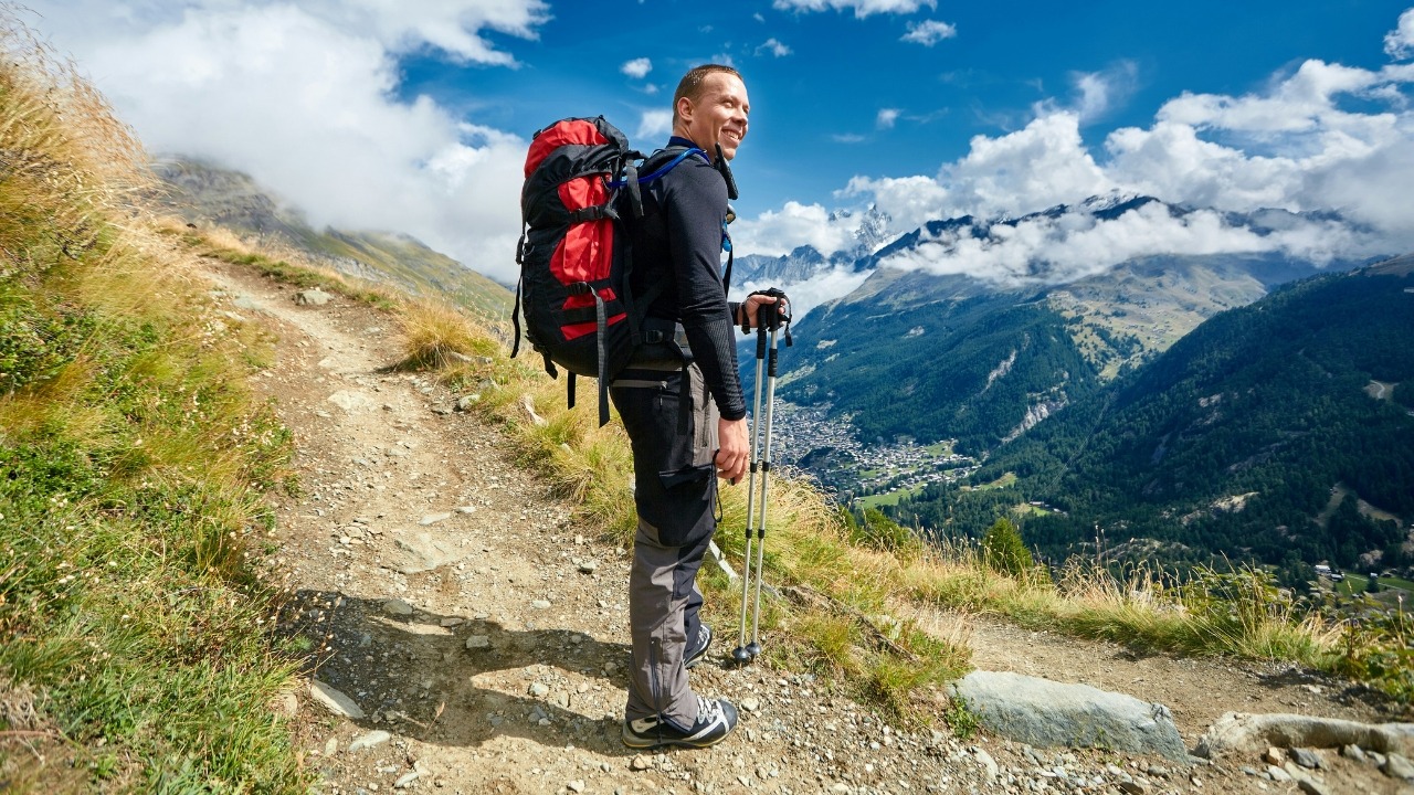 A hiker with a large red backpack and trekking poles looks out over a valley.