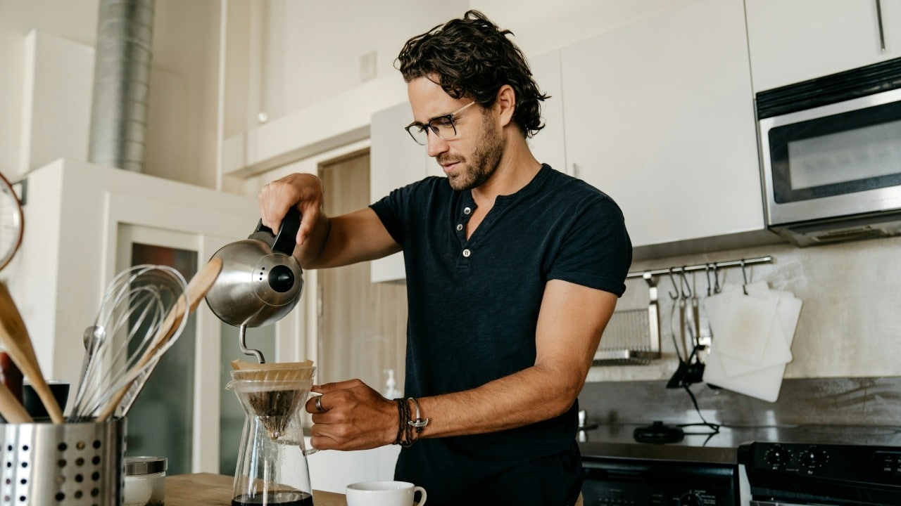 A man in a kitchen pours water from a kettle into a glass coffee carafe.
