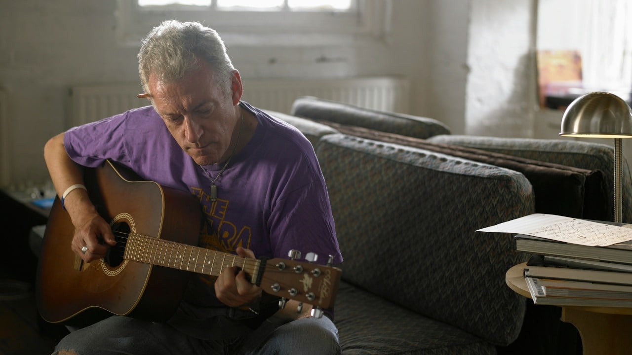 A man in a purple t-shirt plays an acoustic guitar while sitting on a patterned sofa.